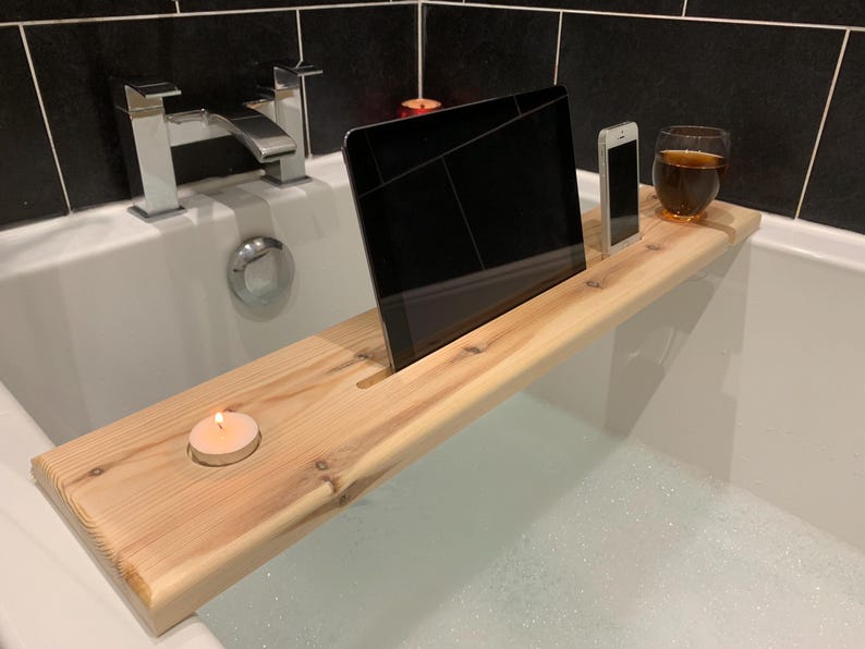 May include: A wooden bath tray with a slot for a tablet, a phone holder, and a space for a candle. The tray is in a bathtub filled with bubbles.