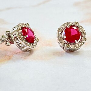 May include: A pair of stud earrings featuring a vibrant red gemstone surrounded by a halo of smaller, clear stones. The earrings have a silver-colored metal setting with intricate detailing around the central stone. The background is a soft, marble-like surface.