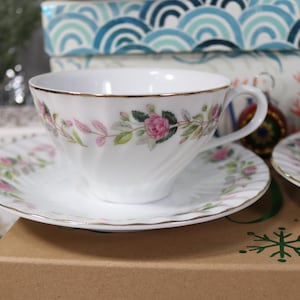 May include: White teacups and saucers with a floral design and gold trim. The teacups and saucers are stacked on top of decorative boxes. The teacups have a pink rose and leaf pattern.