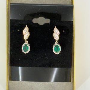 May include: A pair of gold drop earrings featuring oval-cut green gemstones surrounded by small clear stones. The upper portion of each earring has a curved design set with clear stones. The earrings are displayed on a black card in a gold box.