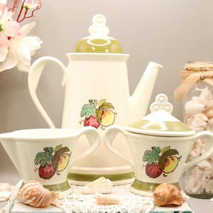 May include: A vintage cream-colored tea set with a teapot, creamer, and sugar bowl. Each piece features a green and red fruit design. The teapot has a matching lid and a curved handle. The sugar bowl has a lid and two handles.