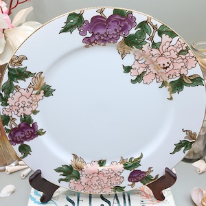 May include: White ceramic plate with a gold rim, adorned with pink and purple floral designs and green leaves. The plate is displayed on a wooden stand, showcasing its decorative pattern.