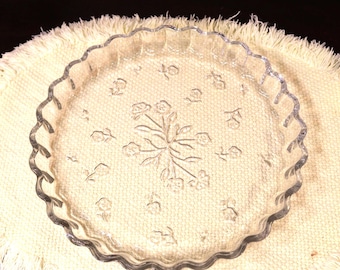 May include: A clear glass pie dish with a scalloped edge and a textured floral pattern embossed on the bottom. The center features a cluster of flowers, while smaller individual floral motifs are arranged in a circular pattern around the base.