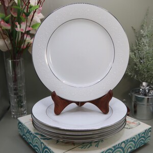 May include: A stack of white ceramic plates with a silver rim and a delicate floral pattern around the edge. The plates are displayed on a wooden stand, with a single plate on top. The plates are part of a dinnerware set.