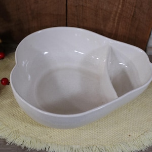 May include: A white ceramic serving dish shaped like a pear with a divided section. The dish is on a light yellow placemat with fringe.