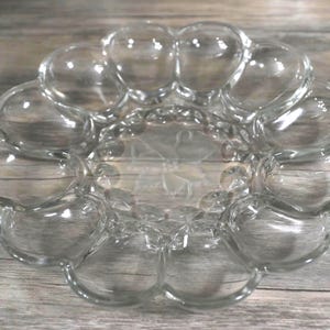 May include: Clear glass serving dish with a flower-like design. The dish has a central area with a floral pattern and is surrounded by bubble-shaped compartments. The glass is transparent, allowing light to pass through.