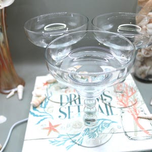 May include: A clear glass champagne serving piece with three bowls, filled with liquid. The stem is clear and has a decorative design. The piece sits on a book with the words "DREAMS SET SAIL" and a seashell theme. A vase and a jar filled with seashells are in the background.