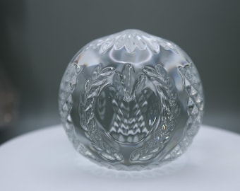 May include: A clear, spherical glass paperweight featuring intricate, etched detailing. The surface displays a central laurel wreath motif surrounding a textured, fan-like pattern, with diamond-cut cross-hatching along the sides.