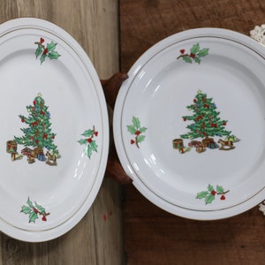 May include: Two white ceramic Christmas plates with a gold rim. Each plate features a green Christmas tree with red and gold ornaments, presents, and a rocking horse. The plates are decorated with green holly leaves and red berries.