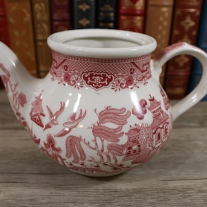 May include: A white teapot with a red and white floral pattern. The teapot has a round body and a spout on one side. The handle is on the opposite side of the spout.