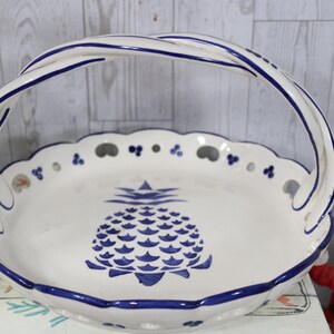 May include: White ceramic serving basket with a blue pineapple design. The basket has a scalloped edge and a twisted handle, both trimmed in blue. A decorative and functional piece.