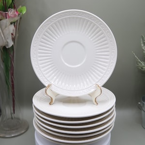 May include: A stack of white ceramic saucers with a detailed, ridged design. The saucers are displayed on a white surface, with one saucer elevated on a decorative stand. The design includes a central indentation for a teacup.