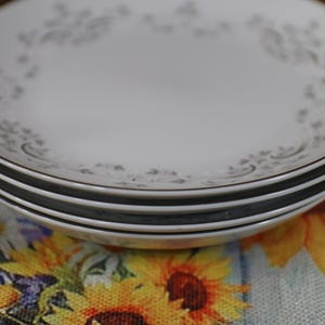 May include: Three white ceramic plates with a delicate floral pattern in grey. The plates are stacked on top of each other.