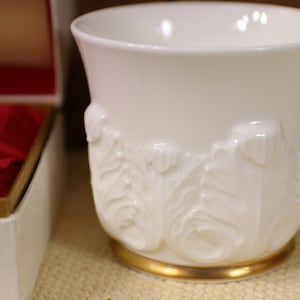 May include: A white ceramic cup with a gold rim and an embossed floral design. The cup is sitting on a beige surface.