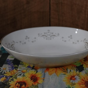 May include: A white ceramic bowl with a silver floral design. The bowl is sitting on a checkered tablecloth with a floral pattern.