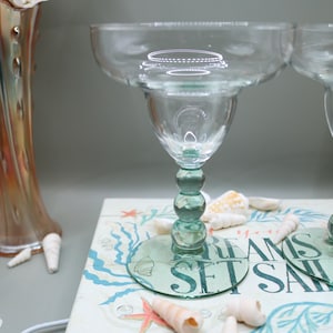 May include: Two clear margarita glasses with teal green stems and bases. The glasses sit on a book with the words "DREAMS SET SAIL". Seashells and a decorative vase are in the background.