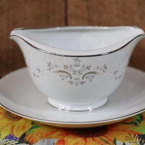 May include: White porcelain gravy boat with a silver trim and a delicate floral design. The boat is sitting on a matching saucer.