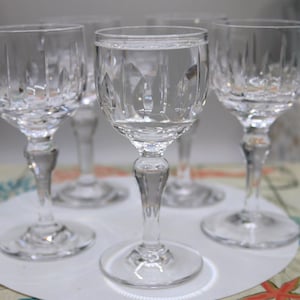 May include: A set of five clear crystal wine glasses. Each glass features a round base, a slender stem, and a bowl with vertical cut patterns. The glasses are arranged on a white surface, showcasing their elegant design.