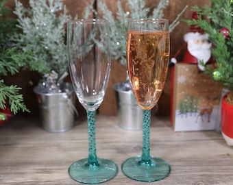 May include: Two champagne flutes with teal stems. One flute is filled with a light pink sparkling beverage.
