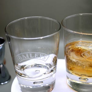 May include: Two clear, lowball glasses, one with the "Baileys" logo etched on it. One glass contains a golden liquid and ice. A metal jigger and bar spoon are in the background, suggesting a cocktail theme.