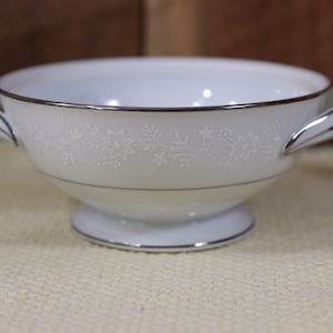 May include: White porcelain soup bowl with two handles and a floral design. The bowl has a silver rim and a silver band around the base.