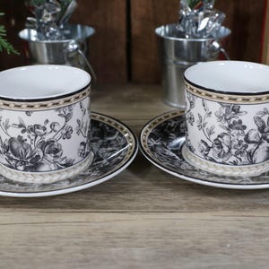 May include: Two white teacups and saucers with black and gold floral patterns. The cups have a gold band around the rim and a gold trim around the base. The saucers have a black and gold floral pattern on the outside and a white center.