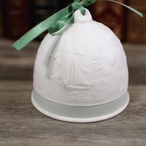 May include: White ceramic bell ornament with a sailboat and sun design. The bell has a green ribbon for hanging. The bell is sitting on a wooden surface.