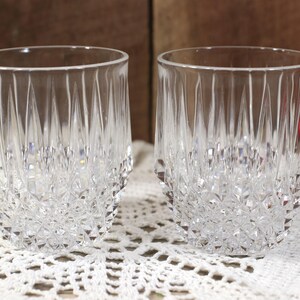May include: Two clear, cut-crystal drinking glasses. The glasses have a diamond-cut pattern on the lower half and vertical cuts on the upper half. They sit on a white, lace doily against a brown wooden background.