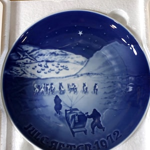 May include: A blue and white decorative plate featuring a winter scene with figures pulling a sled. The plate has a dark blue border and the text "JULEAFTEN 1972". The plate is in a white box.