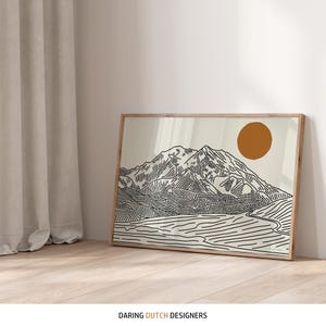 May include: A framed art print depicting a minimalist mountain scene with a rising sun. The artwork uses black lines on a cream background, set within a natural wood frame. The print is leaning against a white wall on a wooden floor.