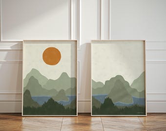 May include: A set of two framed wall art prints featuring a landscape illustration. The left print displays a large orange sun above layered sage green mountains, a blue lake, and a dark green forest. The right print shows similar mountains.