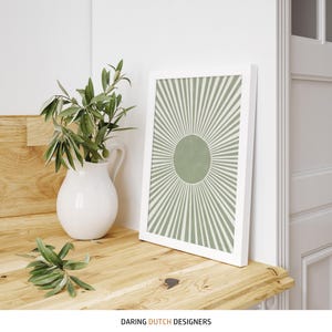 Puede incluir: Una impresi&oacute;n enmarcada en blanco con un dise&ntilde;o de rayos de sol verde y blanco. La impresi&oacute;n est&aacute; sobre una superficie de madera junto a una jarra blanca con hojas verdes.