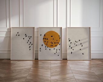 May include: Three framed prints with a minimalist design featuring a flock of birds flying around a yellow sun. The prints are displayed on a light wood floor against a white wall.