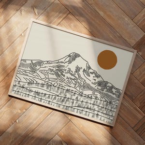 Op de afbeelding: Een ingelijste kunstdruk met een berglandschap en een zon. Het kunstwerk is een minimalistische lijntekening in zwart op een crèmekleurige achtergrond. De lijst is licht houtkleurig en de zon is een effen oranje cirkel.