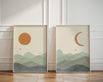 May include: Two framed wall art prints featuring layered mountain landscapes in shades of sage green and dark grey. One print displays a solid orange sun, while the other shows an orange crescent moon. Small black birds are scattered across the sky.