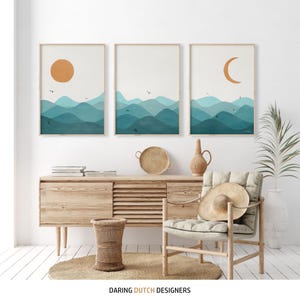 May include: Three abstract landscape prints with a minimalist design featuring a sun, moon, and blue and green mountains. The prints are framed and hung on a white wall above a wooden cabinet.
