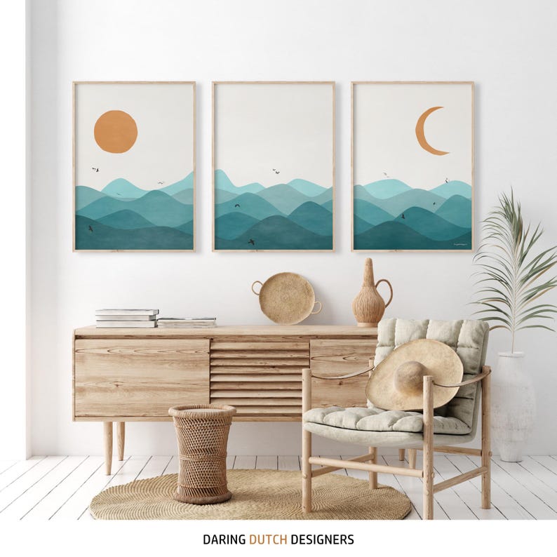 May include: Three abstract landscape prints with a minimalist design featuring a sun, moon, and blue and green mountains. The prints are framed and hung on a white wall above a wooden cabinet.