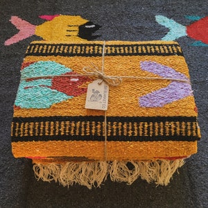 May include: A folded, rectangular blanket with a vibrant yellow, black, red, turquoise, and purple design. The blanket is tied with twine and has a tag that reads "SW BLANKET CO." The blanket has a fringe along the bottom edge. The blanket is on a dark gray surface.