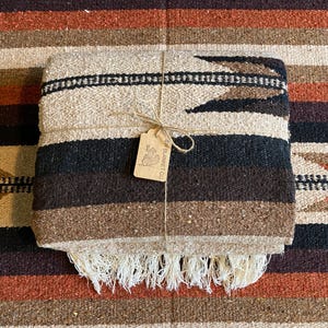 May include: A folded woven blanket featuring horizontal stripes in shades of cream, dark brown, and black. The blanket is tied with twine and includes a rectangular cardboard tag with the text SW BLANKET CO and a cactus illustration.