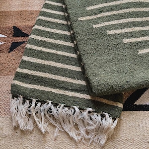 May include: A green and white striped rug with fringe. The rug has a woven texture and is made of natural fibers.