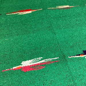 Op de afbeelding: Een levendig groen geweven tapijt met een reeks abstracte, horizontale motieven. Elk motief is samengesteld uit kleine, getextureerde segmenten in tinten wit, felrood en donkerblauw, verspreid over het effen groene achtergrondoppervlak.
