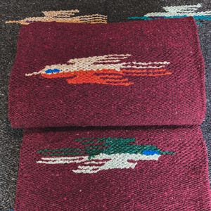 May include: A folded deep burgundy woven blanket featuring two distinct bird motifs. The top bird is rendered in cream and orange with a blue eye detail, while the bottom bird is depicted in green and cream with a blue eye detail.