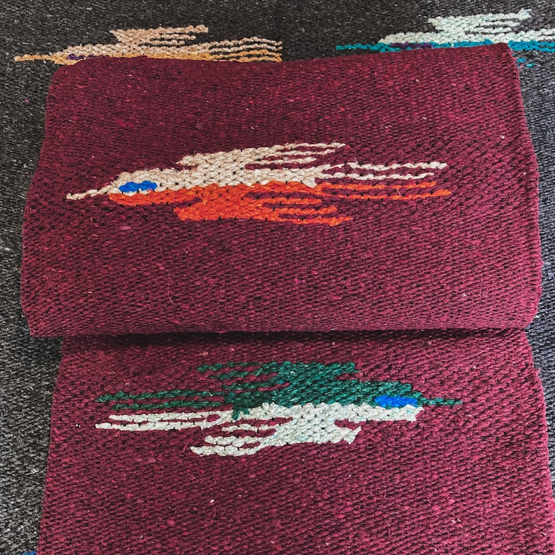 May include: A folded deep burgundy woven blanket featuring two distinct bird motifs. The top bird is rendered in cream and orange with a blue eye detail, while the bottom bird is depicted in green and cream with a blue eye detail.