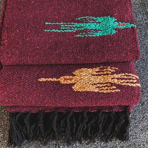 May include: A folded deep burgundy woven blanket featuring a textured surface with a repeating bird motif. The top bird is turquoise, the bottom bird is gold, and the bottom edge of the blanket is finished with a row of black fringe tassels.