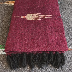 May include: A deep burgundy woven blanket featuring a central beige bird motif with a small red accent. The textile has a textured, heavy-duty weave and is finished with a row of thick, black fringe along the bottom edge of the fabric.