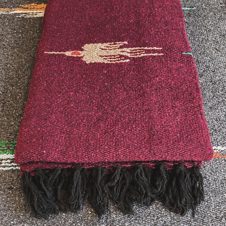 May include: A deep burgundy woven blanket featuring a central beige bird motif with a small red accent. The textile has a textured, heavy-duty weave and is finished with a row of thick, black fringe along the bottom edge of the fabric.