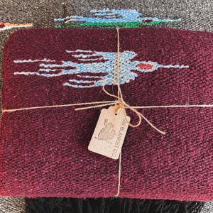 May include: A deep burgundy woven blanket featuring a light blue and white bird pattern. The blanket is tied with brown twine and includes a rectangular tan paper tag printed with a cactus graphic and the text SW BLANKET CO.