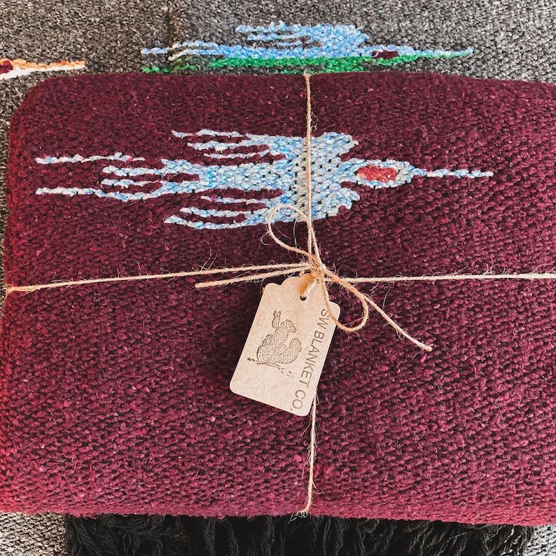 May include: A deep burgundy woven blanket featuring a light blue and white bird pattern. The blanket is tied with brown twine and includes a rectangular tan paper tag printed with a cactus graphic and the text SW BLANKET CO.
