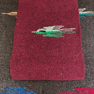 May include: A woven textile featuring a deep burgundy base with a textured, speckled appearance. Centered on the fabric is a stylized bird graphic rendered in shades of light green and dark green, with thin horizontal lines extending from the wings.