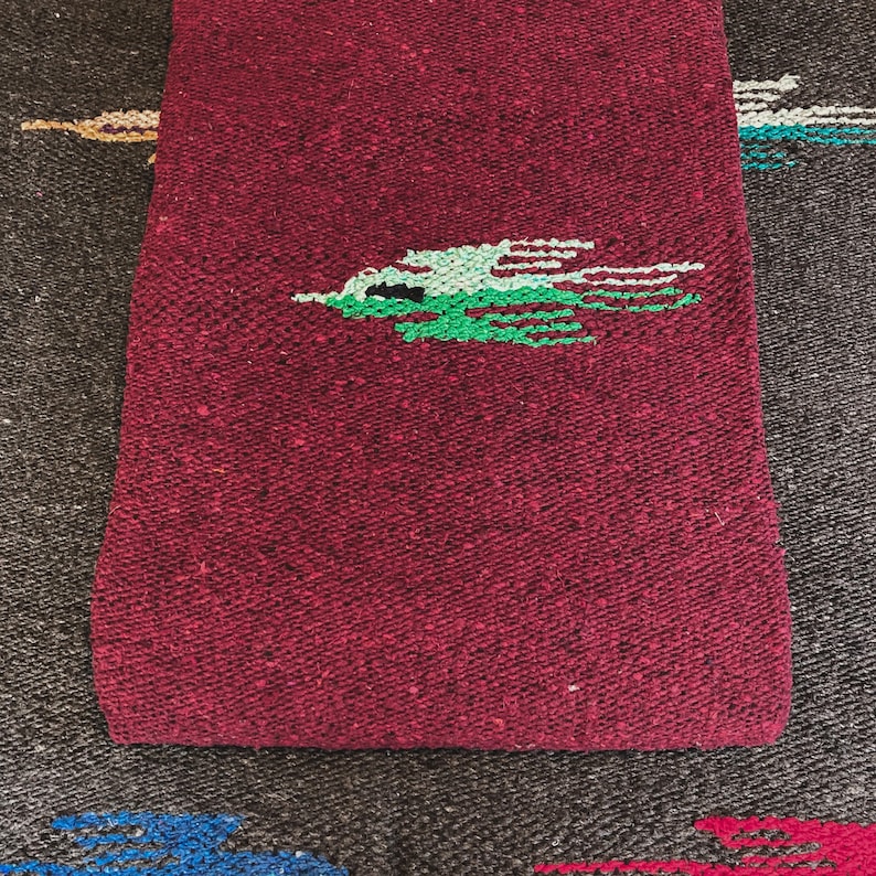 May include: A woven textile featuring a deep burgundy base with a textured, speckled appearance. Centered on the fabric is a stylized bird graphic rendered in shades of light green and dark green, with thin horizontal lines extending from the wings.